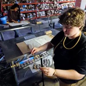 A young man holds a tube of glass above a flame while a young woman works on a neon pattern.