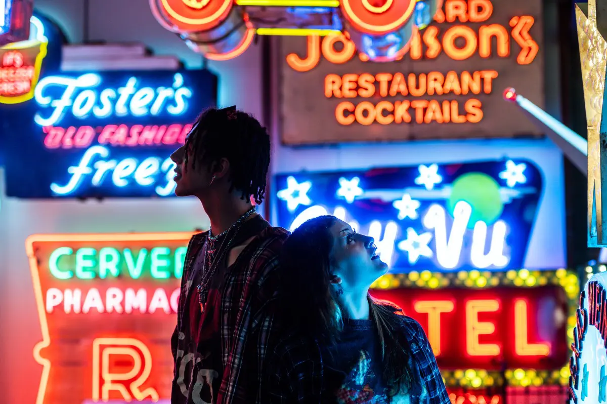 A young couple surrounded by vibrant neon signs looks up in awe